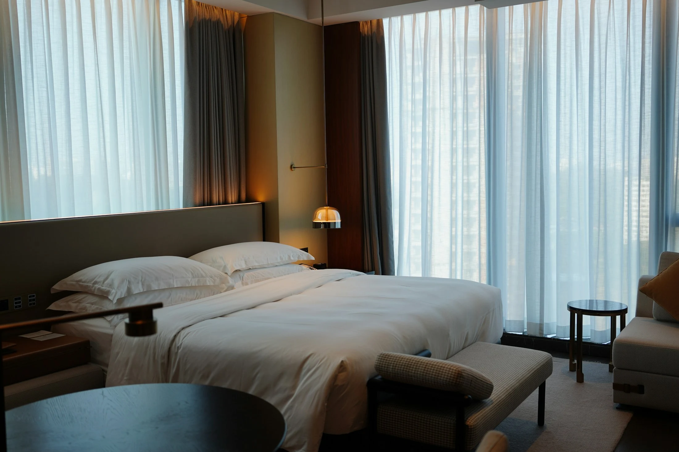 Lichtdurchflutetes Hotelzimmer mit weißen Betttextilien, warmen Materialien und großen Fenstern